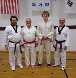 Four men in white taekwondo uniforms and belts. Two are wearing red belts and are standing between two black belt instructors.